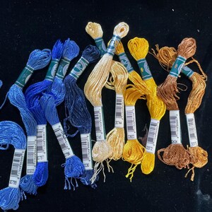 May include: A collection of embroidery floss in various colors, including shades of blue, yellow, and brown. Each skein is labeled with a product number and wrapped with a green and white band. The floss is arranged on a dark surface.
