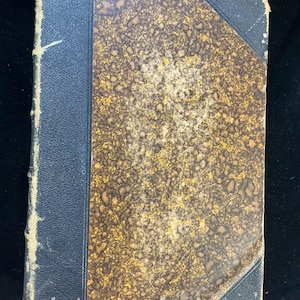 May include: An antique book with a worn, textured cover. The cover features a mottled pattern in shades of brown and gold, with a dark blue spine and corner accents. The edges show signs of age and wear.