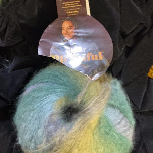 May include: A ball of multi-colored yarn in shades of green, blue, and yellow. The yarn is fluffy and soft, with a slight sheen. A partially visible label shows a woman and the word "Beautiful".