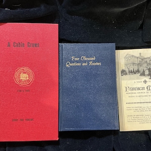May include: Three vintage books are displayed. The first book is red with gold lettering and a circular emblem, titled "A Cabin Grows." The second book is blue with gold lettering, titled "Four Thousand Questions and Answers." The third is a pamphlet.