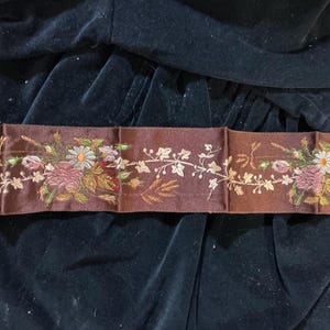 May include: A brown embroidered ribbon with floral and leaf designs. The ribbon features a repeating pattern of flowers in shades of pink, white, and yellow, with green and brown leaves. The background is a dark, velvety fabric.
