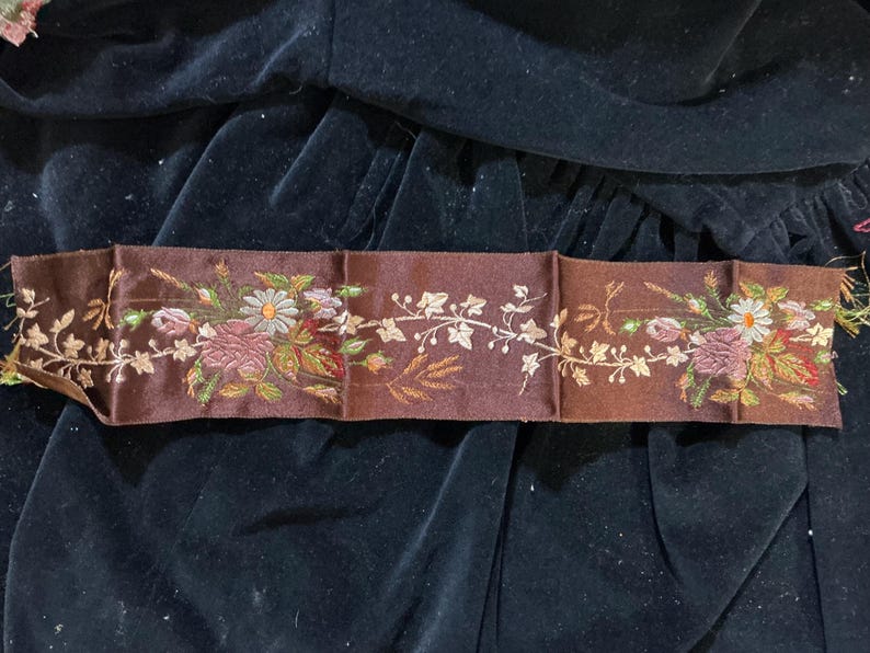 May include: A brown embroidered ribbon with floral and leaf designs. The ribbon features a repeating pattern of flowers in shades of pink, white, and yellow, with green and brown leaves. The background is a dark, velvety fabric.