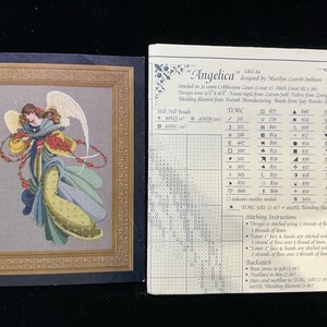 May include: A cross-stitch pattern titled "Angelica" featuring a detailed image of an angel with flowing hair, wings, and a flowing gown. The pattern includes a color chart and stitching instructions. The design is framed in a gold border.