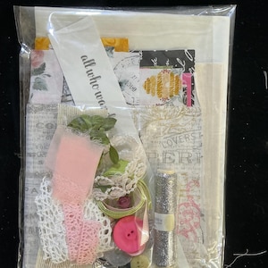 May include: A clear plastic bag filled with craft supplies. The bag contains various items, including pink lace, buttons, ribbon, and a spool of silver glitter thread. A white tag with the text "all who wander" is also visible.