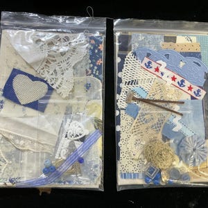May include: Two clear plastic bags filled with craft supplies. One bag contains white lace, blue fabric scraps, and a blue heart. The other bag has blue and white fabric, lace, and nautical-themed embellishments.