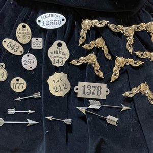 Assortment of 10 metal number tags and 6 flat backed arrows and 8 scroll corner pieces in brass. Maybebe Tim Holtz. In plastic box.