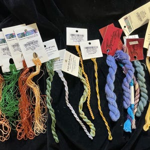May include: A collection of colorful embroidery floss skeins in shades of orange, green, yellow, blue, and purple. Each skein is attached to a tag with product information. The floss is arranged on a dark surface, showcasing the vibrant colors.