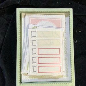 May include: A light green rectangular box filled with various stationery items. The box contains stacks of white paper, some with red-bordered labels, and other sheets with decorative patterns. The box is set against a dark background.
