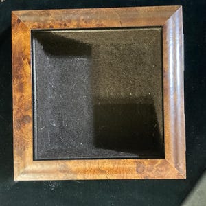 May include: Square wooden picture frame with a dark interior. The frame has a warm, brown finish with a natural wood grain pattern. The frame is empty, ready to display a photograph or artwork. The frame is sitting on a dark surface.