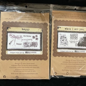 May include: Two clear plastic packages containing rubber stamps. The first package features a stamp with the text "happy birthday you make my heart sing happy" and a dragonfly. The second package features a stamp with the text "when I see you. I automatically smile when I see you." and a grid of circles. Both packages have the text "Designed with love by" and the company logo.