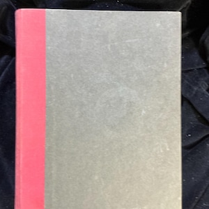 May include: A hardcover book with a gray cover and a red spine. The book is rectangular and appears to be closed. The cover has a slightly textured appearance.
