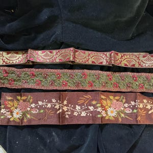 May include: Three decorative ribbons with intricate designs. The top ribbon is red and gold with a paisley pattern. The middle ribbon is green and red with floral embroidery. The bottom ribbon is brown with floral and leaf embroidery.
