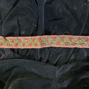 May include: A decorative trim featuring a floral pattern with red and green embroidered flowers. The trim has a pink border and is embellished with small, shiny accents. The trim is placed on a black velvet fabric.