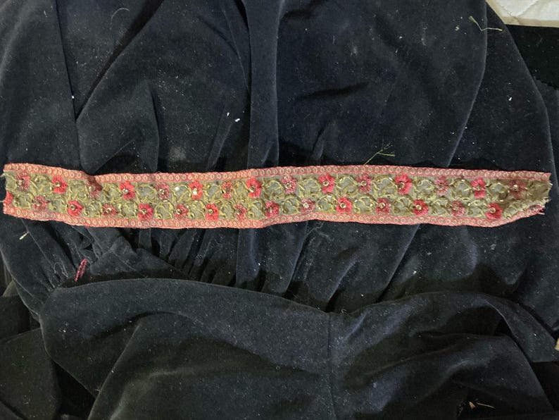 May include: A decorative trim featuring a floral pattern with red and green embroidered flowers. The trim has a pink border and is embellished with small, shiny accents. The trim is placed on a black velvet fabric.