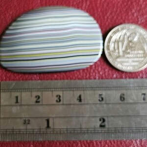 Op de afbeelding: Een close-up van een veelkleurige agaat steen met een gestreept patroon. De steen is ongeveer 5 cm in diameter en ligt op een rood oppervlak naast een liniaal.