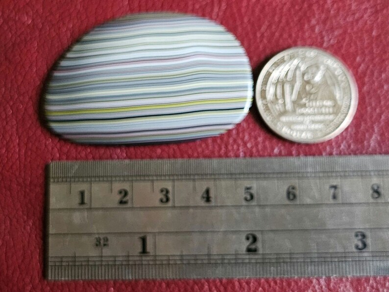 Op de afbeelding: Een close-up van een veelkleurige agaat steen met een gestreept patroon. De steen is ongeveer 5 cm in diameter en ligt op een rood oppervlak naast een liniaal.