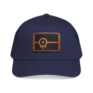 Puede incluir: Gorra de béisbol azul marino con un parche de cuero marrón. El parche presenta un rectángulo negro con una estrella y un diseño de goteo, y una línea horizontal marrón. La gorra tiene una visera curva y un ajuste estructurado.