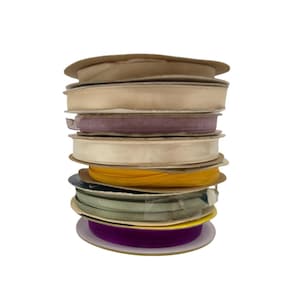 May include: A stack of spools of ribbon in various colors, including beige, purple, yellow, and green. The ribbons are made of a silky material and are wound around cardboard spools. The stack is on a white background.