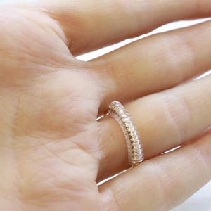 May include: A silver ring with a textured band. The ring is worn on a finger.