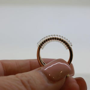 May include: A gold-coloured ring with a clear, beaded band. The ring is held by a person. The beads are small and evenly spaced, creating a textured appearance. The ring's band is smooth and shiny.