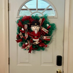 May include: A Christmas wreath featuring a nutcracker figure in a red uniform and black hat. The wreath is made of green mesh, red ribbons, and decorative ornaments, hanging on a white door.