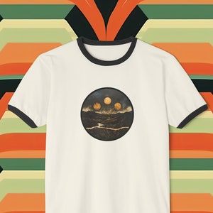 Puede incluir: Camiseta blanca con cuello de canalé negro y un gráfico circular de un paisaje con tres lunas.