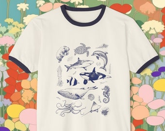 May include: A white t-shirt with a navy blue ringer. The shirt features a blue and white illustration of various sea creatures, including a whale, a seahorse, a shark, a turtle, and an octopus.