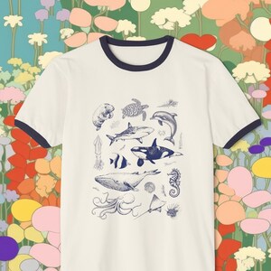 May include: A white t-shirt with a navy blue ringer. The shirt features a blue and white illustration of various sea creatures, including a whale, a seahorse, a shark, a turtle, and an octopus.
