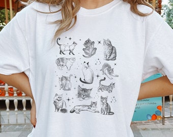 Może przedstawiać: Biały t-shirt z czarno-białym nadrukiem graficznym przedstawiającym różne koty w różnych pozach. Grafika jest umieszczona centralnie na koszulce i przedstawia różne rasy kotów, w tym kota pręgowanego, trójkolorowego i syjamskiego.