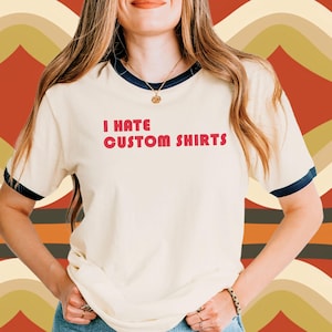 Puede incluir: Camiseta color crema con ribetes azul marino en el cuello y las mangas. La camiseta tiene el texto "I HATE CUSTOM SHIRTS" en letras rojas. La persona de la imagen lleva vaqueros azules.