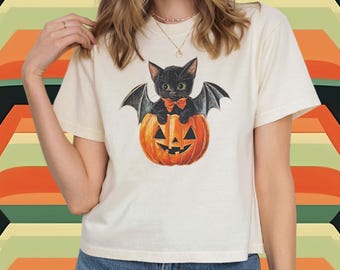May include: Ivory-colored t-shirt featuring a Halloween-themed graphic. The design showcases a black cat with bat wings, wearing a bow tie, and sitting inside a carved orange pumpkin. The shirt is paired with blue jeans.