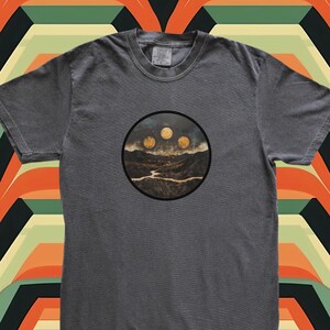 Op de afbeelding: Een donkergrijs T-shirt met een ronde afbeelding van een landschap met drie manen aan de hemel. De manen zijn lichtgeel van kleur.