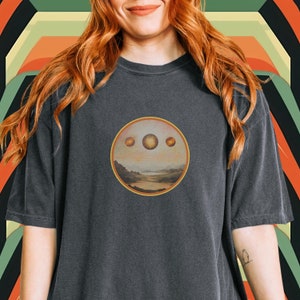 Puede incluir: Una camiseta gris oscuro con un gráfico circular de un paisaje con tres soles. Los soles son naranja y amarillo con un efecto de estrella.