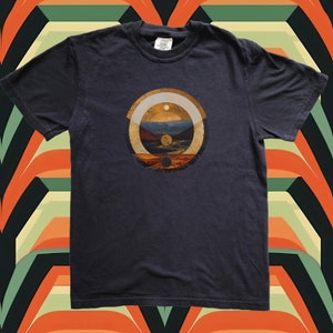 Puede incluir: Una camiseta gris oscuro con un diseño gráfico circular que presenta un paisaje estilizado con una puesta de sol, montañas y un río. El gráfico está en tonos de marrón, naranja, amarillo y blanco.