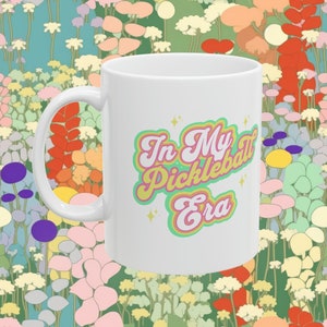 May include: White ceramic coffee mug with a green, pink, and yellow design that says "In My Pickleball Era".