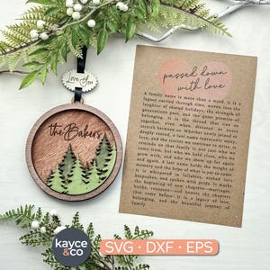 Könnte beinhalten: Hölzernes Familiennamensornament mit einer Waldszene und dem Text "the Bakers". Enthält eine Karte mit dem Text "passed down with love". Das Ornament ist rund mit einem dunklen Holzrahmen und einem schwarzen Band.