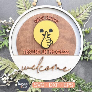 Può includere: Un cartello rotondo in legno con cornice bianca e centro marrone. Il cartello presenta un'emoji gialla con un dito sulla bocca, le parole "QUIET PLEASE" e "TESTING IN PROGRESS". La parola "welcome" è scritta in corsivo in basso.