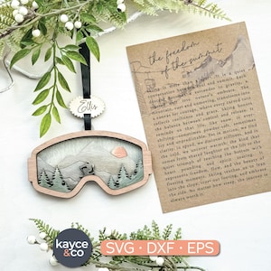 May include: Wooden ski goggles ornament with a mountain scene and a small tag that says "Ellis". The ornament is hanging from a black ribbon. A card with text about skiing is in the background. The ornament is approximately 4 inches wide.
