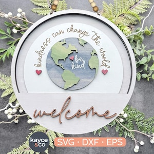 May include: A round wooden sign with the text "kindness can change the world" and a globe graphic with the text "be kind". The sign also says "welcome" in a decorative font. The sign is surrounded by greenery.