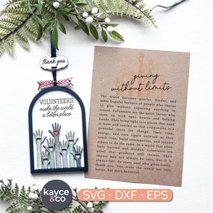 May include: A wooden ornament with the text "VOLUNTEERS make the world a better place" and illustrations of hands. A card next to it reads "giving without limits". The ornament has a black border and a ribbon.