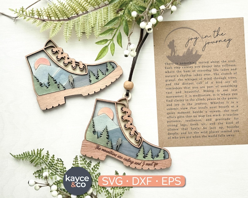 Op de afbeelding: Houten wandelschoenornamenten met een berg- en zonontwerp. De laarzen zijn bruin en blauw met een zwart touwtje om op te hangen. De tekst op de laarzen luidt "The mountains are calling and I must go."