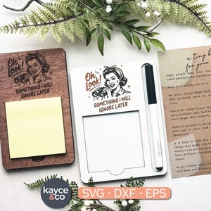 Puede incluir: Dos portamemos de madera, uno marrón y otro blanco, con el texto "Oh Look! Something I Will Ignore Later" y un gráfico de mujer retro. El soporte marrón tiene un bloc de notas adhesivas amarillo. Un bolígrafo está con el soporte blanco. Vegetación y una tarjeta en el fondo.