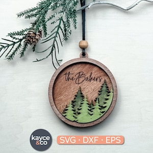 May include: Circular wooden Christmas ornament with the text "the Bakers" above a forest scene with green trees. The ornament hangs from a black cord with a wooden bead. The design is laser cut.