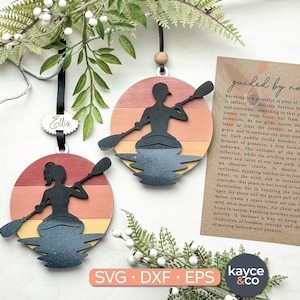 May include: Wooden kayak ornaments with a sunset design. Each ornament features a silhouette of a person kayaking on a lake. The ornaments are round, with a black ribbon for hanging. The text "SVG DXF EPS" is visible.