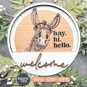 Könnte beinhalten: Ein runder Willkommensgruß aus Holz mit einer Eselillustration. Das Schild hat einen weißen Rand und einen hellbraunen Holzgrund mit den Worten "hay. hi. hello." und "welcome" in einer Schriftart. Das Schild enthält auch den Text "kayce & co" und "SVG • DXF • EPS".