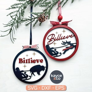 May include: Two Christmas ornaments with the word "Billieve" in red script. One ornament features a person on a sled being pulled by a bison, the other a reindeer. Both have a red and white ribbon.