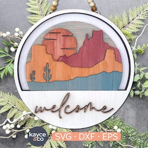 May include: A wooden welcome sign with a layered design of a desert landscape, including mountains, a sunset, and cacti. The sign is painted in shades of red, orange, blue, and brown. The word "welcome" is written in a cursive font below the landscape.
