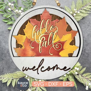 May include: A round wooden sign with a white background and a brown woodgrain border. The sign features a layered design of fall leaves in shades of orange, red, and yellow. The text "hello fall" is written in a decorative script font, and the word "welcome" is written in a cursive font below the leaves.