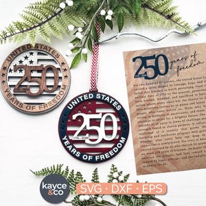 Puede incluir: Dos adornos de madera y una tarjeta que celebra 250 años de libertad. Un adorno es marrón con un diseño de estrellas y rayas, el otro es rojo, blanco y azul. La tarjeta tiene texto sobre el hito.