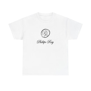 Könnte beinhalten: Ein weißes T-Shirt mit einem schwarzen kreisförmigen Logo mit den Buchstaben "PR" und dem Namen "Phillipe Ray" in einer kursiven Schriftart. Das Shirt besteht aus einem weichen Material und hat einen klassischen Rundhalsausschnitt.